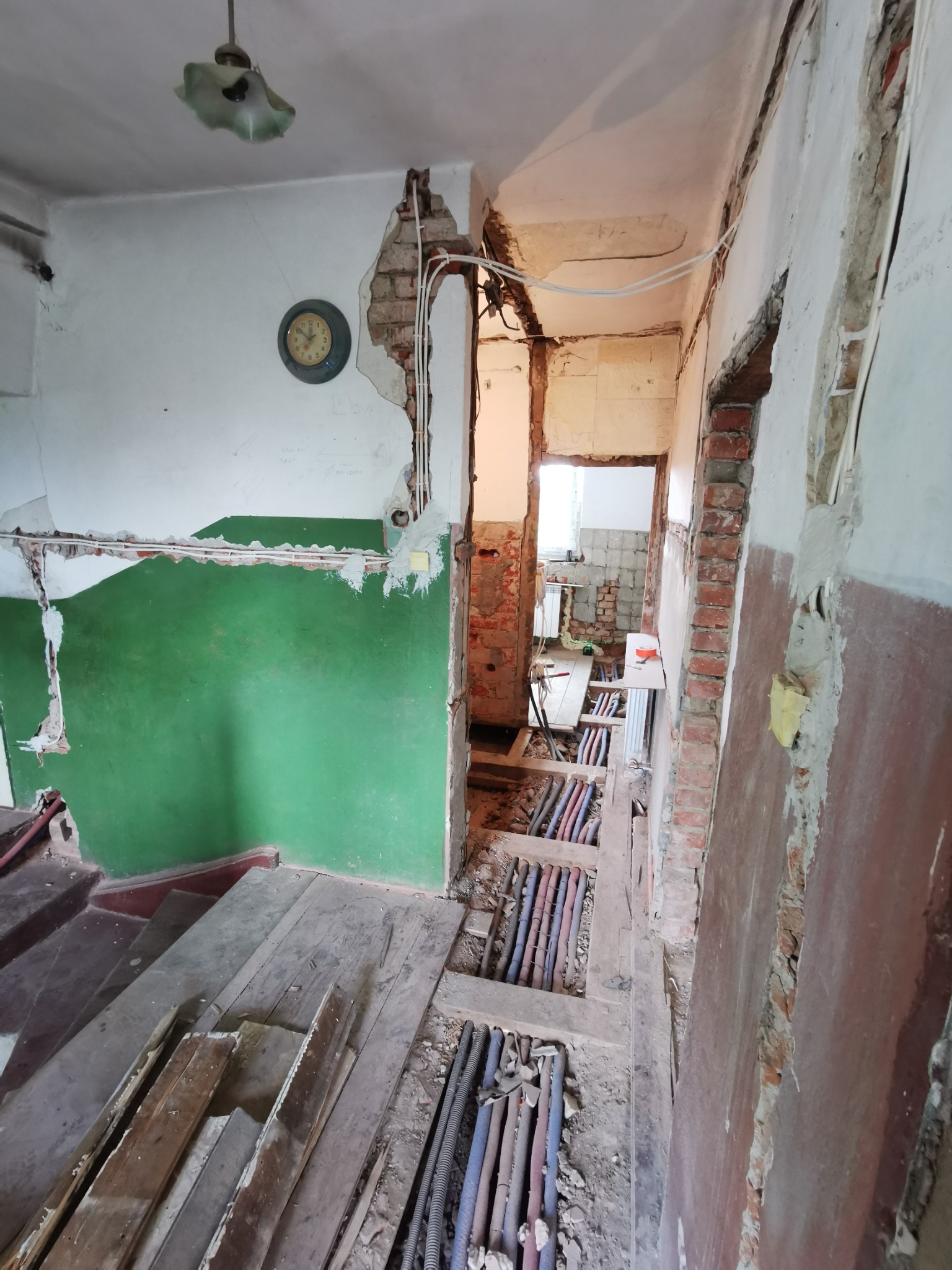 Wnętrze mieszkania w trakcie intensywnego remontu: odsłonięte cegły, fragmenty instalacji elektrycznej i rur grzewczych, stara lampa sufitowa i zegar na ścianie z zielonym fragmentem, widoczne...