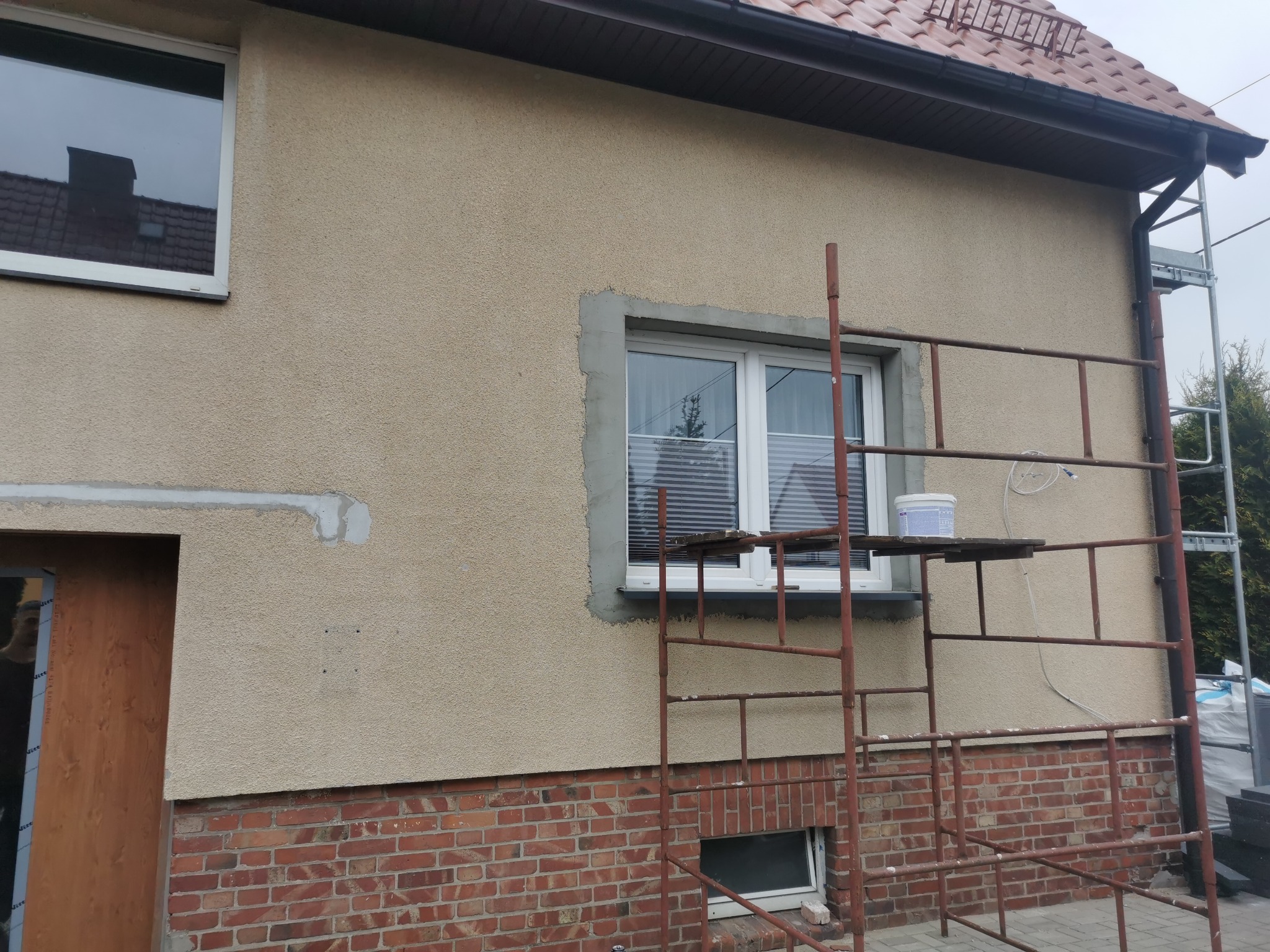 Elewacja domu jednorodzinnego w trakcie ocieplania, widoczne rusztowanie, wiadro z zaprawą na podeście, otynkowane obramowanie okna, ślady po usuniętych elementach na ścianie.