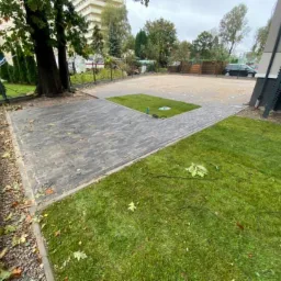 Ukończona nawierzchnia z kostki brukowej w odcieniach szarości, z centralnym elementem trawnika, otoczona zielenią i ogrodzeniem posesji w słoneczny dzień.