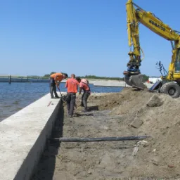 Żółta koparka Komatsu PW160 przy betonowym nabrzeżu, trzech robotników w pomarańczowych koszulkach wyrównuje teren pod instalacje, w tle widoczne jezioro i konstrukcja betonowa na wodzie.
