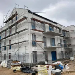 Budynek wielorodzinny w trakcie ocieplania elewacji, z widocznymi rusztowaniami, materiałami izolacyjnymi i stosami zapraw murarskich.