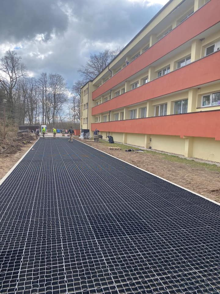 Metalowa kratka drenażowa ułożona wzdłuż budynku, pracownicy w oddali, pochmurne niebo.