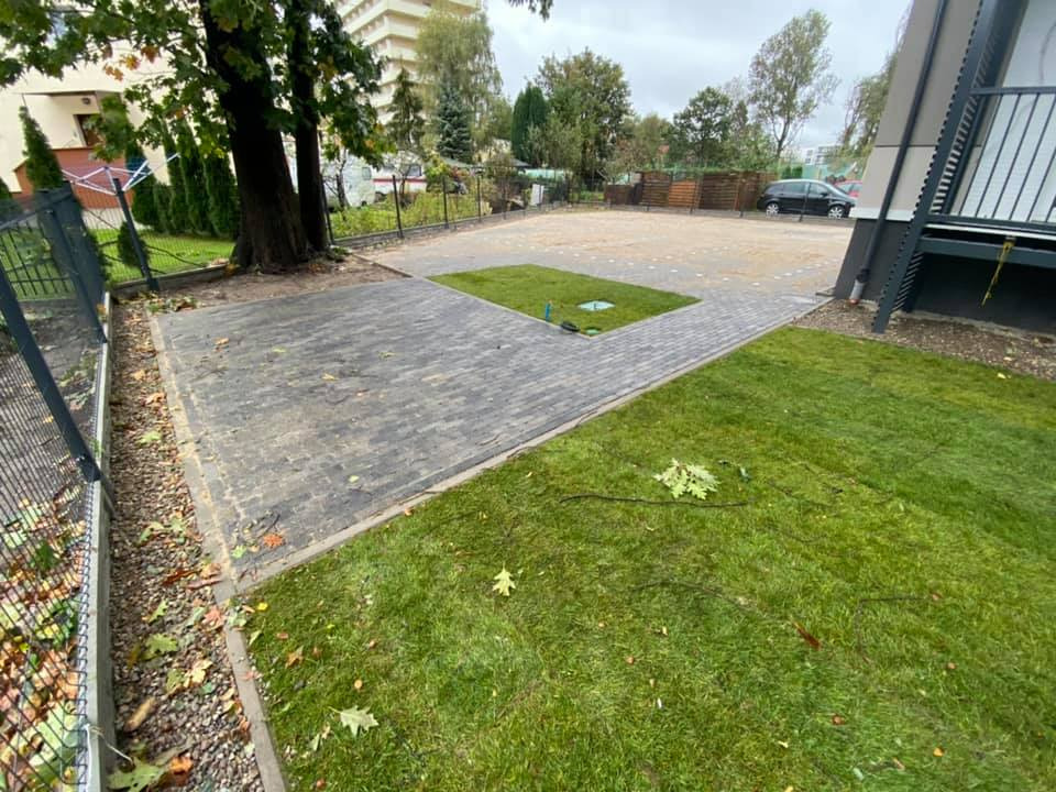 Ukończona nawierzchnia z kostki brukowej w odcieniach szarości, z centralnym elementem trawnika, otoczona zielenią i ogrodzeniem posesji w słoneczny dzień.