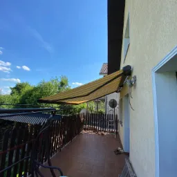 Żółto-biała markiza nad tarasem z brązową podłogą, widok na błękitne niebo i zieleń ogrodu.