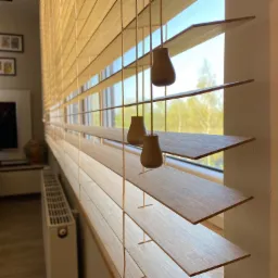 Jasne drewniane żaluzje poziome z widocznymi sznurkami i drewnianymi obciążnikami, częściowo otwarte, w tle widok na zieleń za oknem i fragment wnętrza pokoju z grzejnikiem.