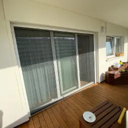 Zastosowanie przesuwnej moskitiery ramkowej w Brzegu na balkonie z drewnianą podłogą, widoczny fragment stołu z talerzem i resztkami jedzenia oraz donice z kwiatami.