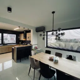 Nowoczesne, jasne wnętrze: otwarta kuchnia z drewnianymi szafkami i czarnymi akcentami, jadalnia z dużym stołem i designerską lampą. Okna przesłonięte poziomymi listwami.