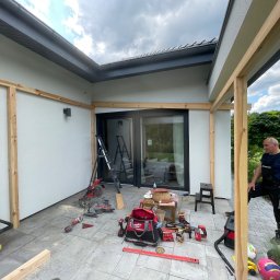 Rolety i Moskitiery Krzysztof Dragan - Montaż drewnianej konstrukcji na tarasie z użyciem narzędzi. Widoczny pracownik w kombinezonie, drabina i sprzęt budowlany na szarej kostce brukowej.