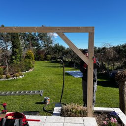 Rolety i Moskitiery Krzysztof Dragan - Montaż drewnianej pergoli w ogrodzie: pracownik w trakcie łączenia elementów konstrukcyjnych na tle zielonego trawnika i błękitnego nieba. Widoczne narzędzia i drabina.