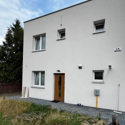 Rolety i Moskitiery Krzysztof Dragan - Nowoczesny, dwukondygnacyjny dom z białymi ramami okiennymi i drewnianymi drzwiami wejściowymi. Widoczne rolety w oknach. Uwagę zwraca minimalistyczny design i otoczenie budynku.