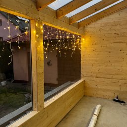 Rolety i Moskitiery Krzysztof Dragan - Drewniana konstrukcja z oknami, udekorowana girlandą świetlną. Widoczne elementy konstrukcyjne i fragment otoczenia zewnętrznego przez okno.