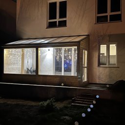 Rolety i Moskitiery Krzysztof Dragan - Wieczorny widok na dobudowany, przeszklony balkon z drewnianą konstrukcją, oświetlony girlandami. Widoczne schody i oświetlenie ogrodowe.