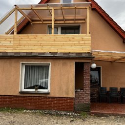 Rolety i Moskitiery Krzysztof Dragan - Dom z drewnianą zabudową balkonu i pergolą z przezroczystym zadaszeniem. Widoczne krzesła na tarasie i ceglana podmurówka. Ujęcie z dołu, pochmurne niebo w tle.