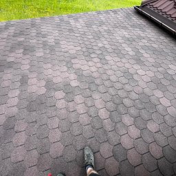 Rolety i Moskitiery Krzysztof Dragan - Dach pokryty brązowo-szarą dachówką bitumiczną w kształcie rybiej łuski, widok z góry, fragment rynny i trawnika w tle, stopa w bucie na dachu.