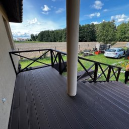 Rolety i Moskitiery Krzysztof Dragan - Nowoczesny taras z deski kompozytowej w kolorze ciemnego brązu, z czarną balustradą i widokiem na ogród. Na tarasie stoi biała kolumna, a w tle widać dom i samochód.
