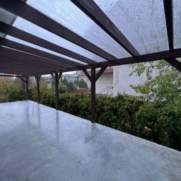 Rolety i Moskitiery Krzysztof Dragan - Zadaszenie tarasu z brązową konstrukcją drewnianą i przezroczystym dachem z tworzywa. Widok na ogród i budynki w tle. Lekko mokra nawierzchnia tarasu.