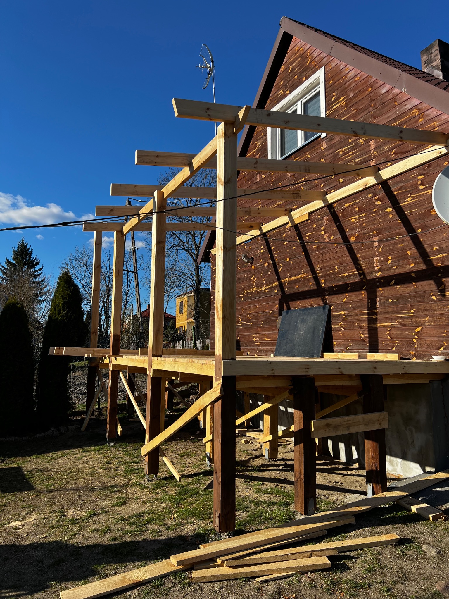 Konstrukcja drewniana tarasu przy brązowym domu z widoczną anteną satelitarną i elementami montażowymi. Jasne drewno kontrastuje z ciemnym tłem.