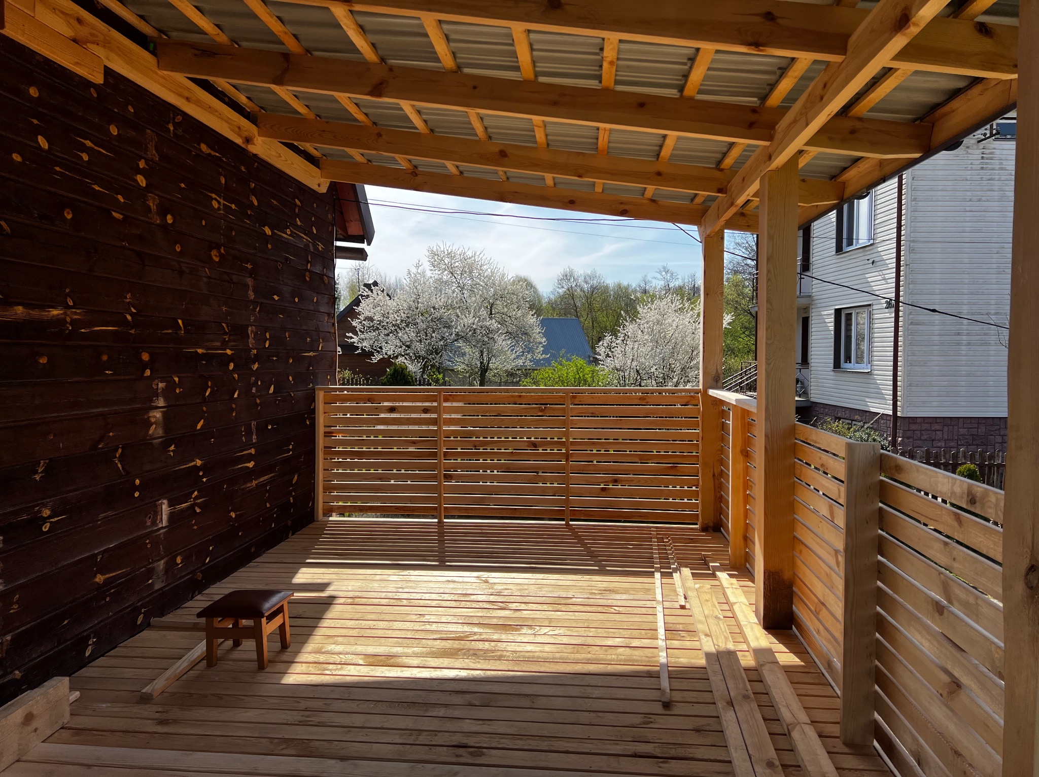 Drewniany taras z zadaszeniem w trakcie budowy, widoczne deski podłogowe, balustrady z poziomych listew i konstrukcja dachu. W tle kwitnące drzewa i budynek.