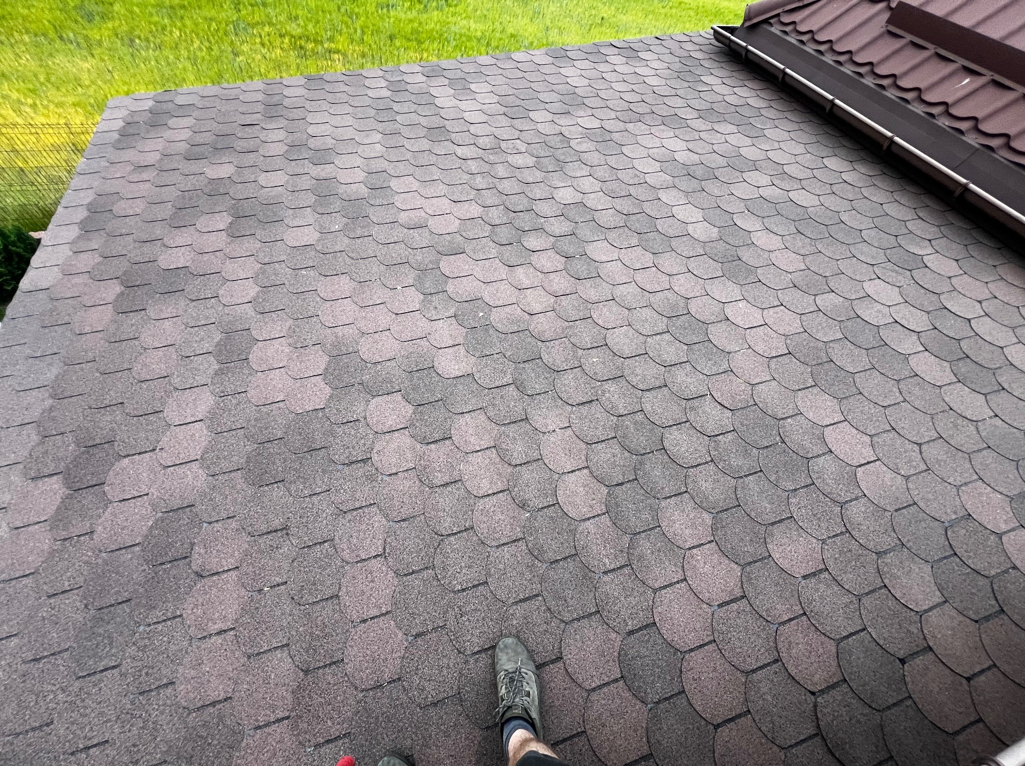 Dach pokryty brązowo-szarą dachówką bitumiczną w kształcie rybiej łuski, widok z góry, fragment rynny i trawnika w tle, stopa w bucie na dachu.