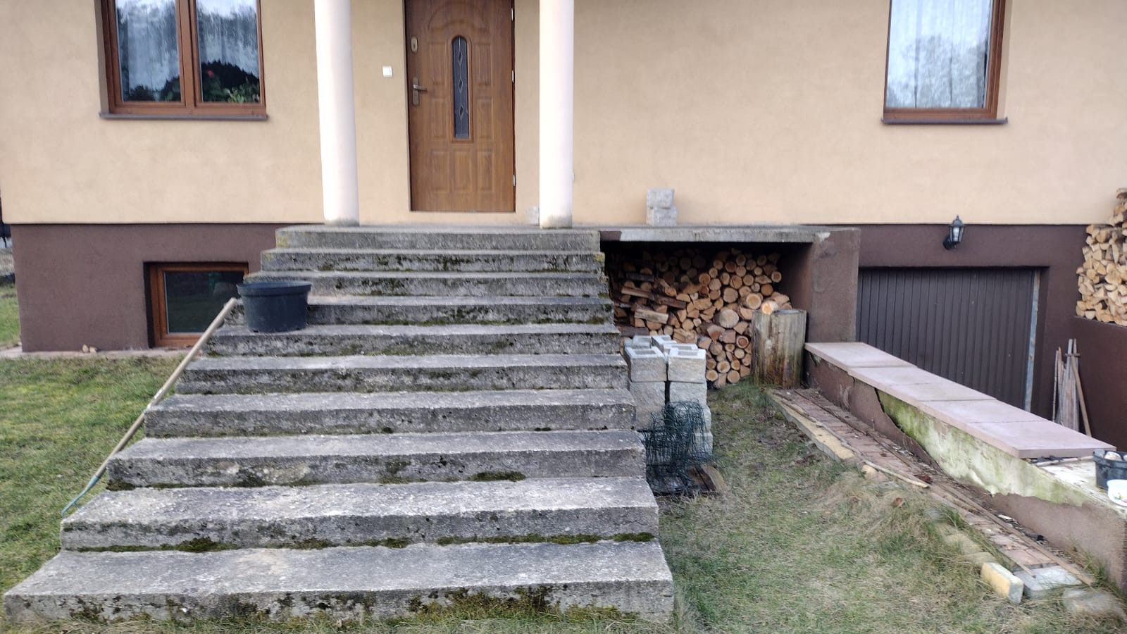 Wejście do domu z betonowymi schodami, widoczne drewno opałowe pod podcieniem. Ujęcie z dołu, podkreślające surowość materiałów i funkcjonalność przestrzeni.