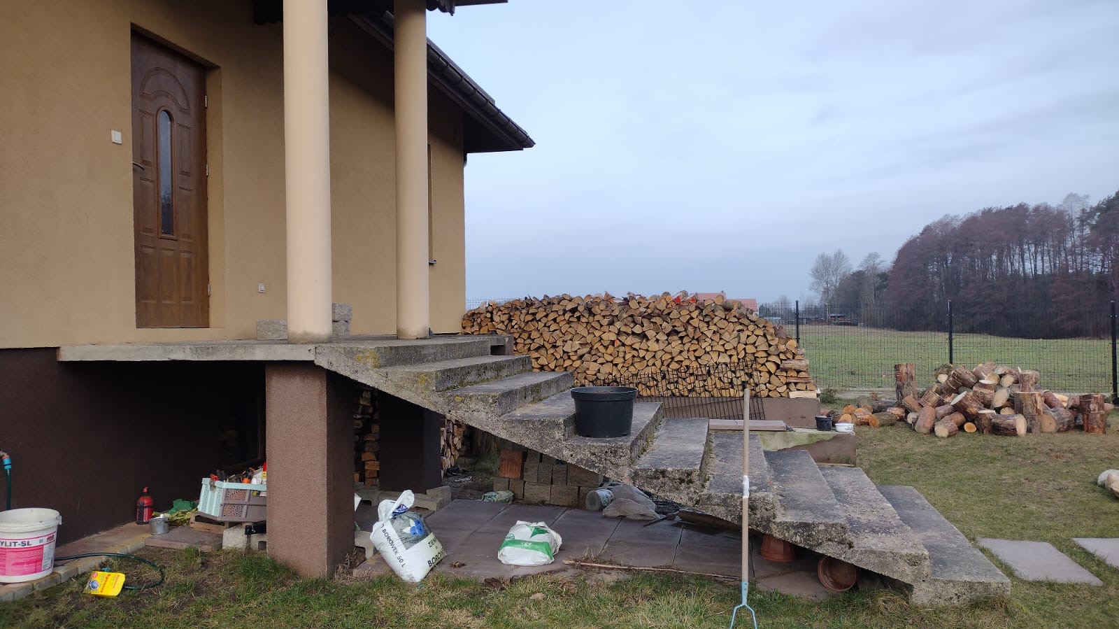 Betonowe schody wejściowe do domu z kolumnami, obok sterta drewna opałowego. Widoczne worki z zaprawą i narzędzia ogrodnicze. Tło: ogrodzenie i las.