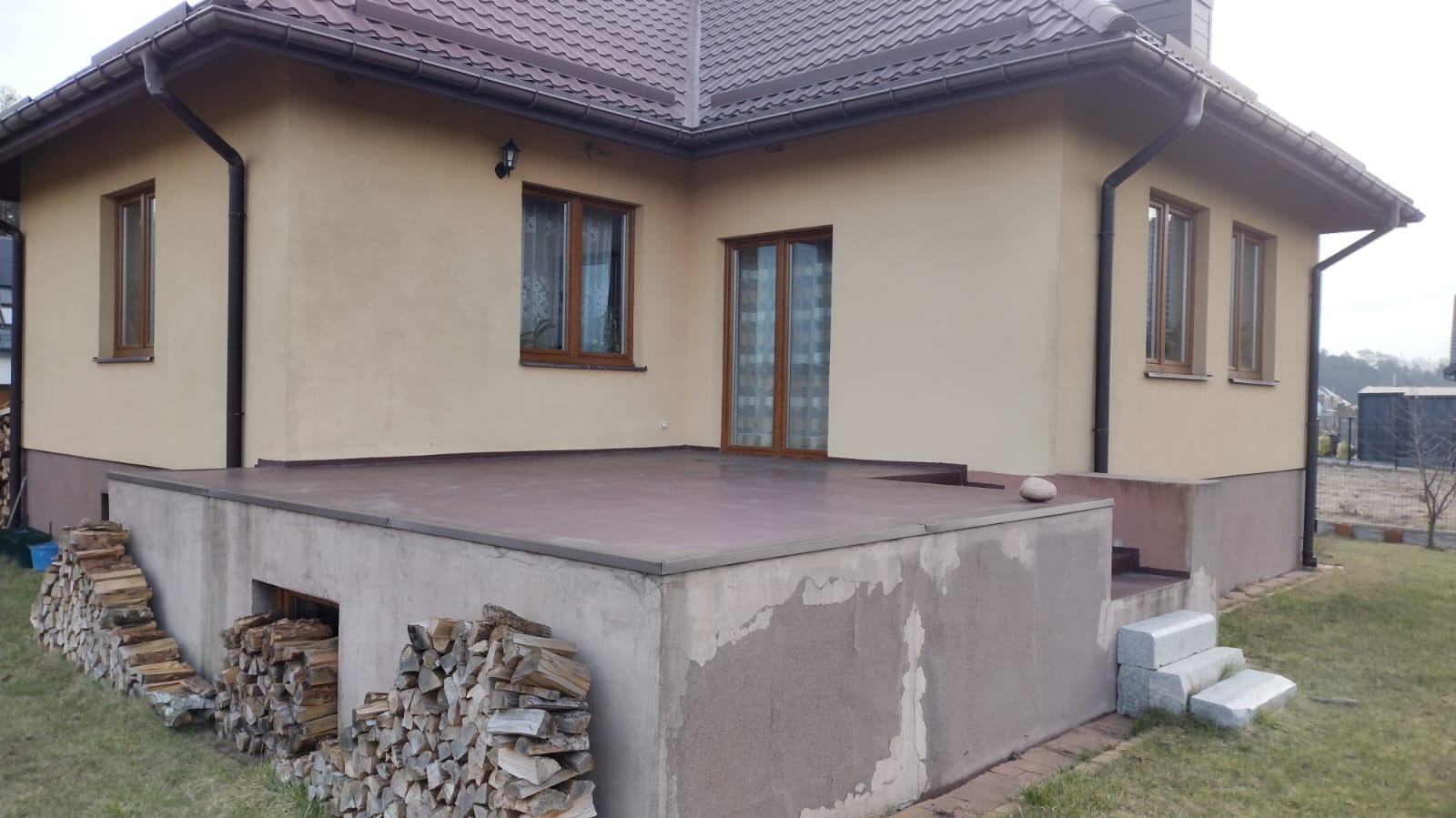 Wykończony taras na podwyższeniu przy domu z brązowym dachem. Na pierwszym planie stos drewna. Widoczne okna z brązowymi ramami i schody z kamienia.