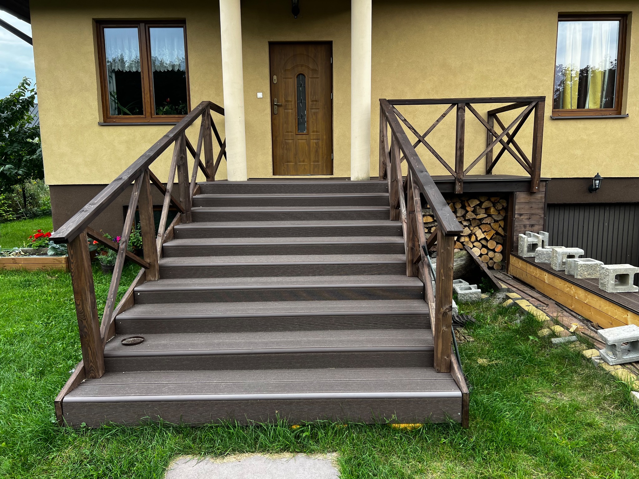 Schody wejściowe z kompozytu, z drewnianą balustradą, prowadzące do domu z żółtą elewacją. Widoczne drewno na opał i bloczki betonowe obok schodów.
