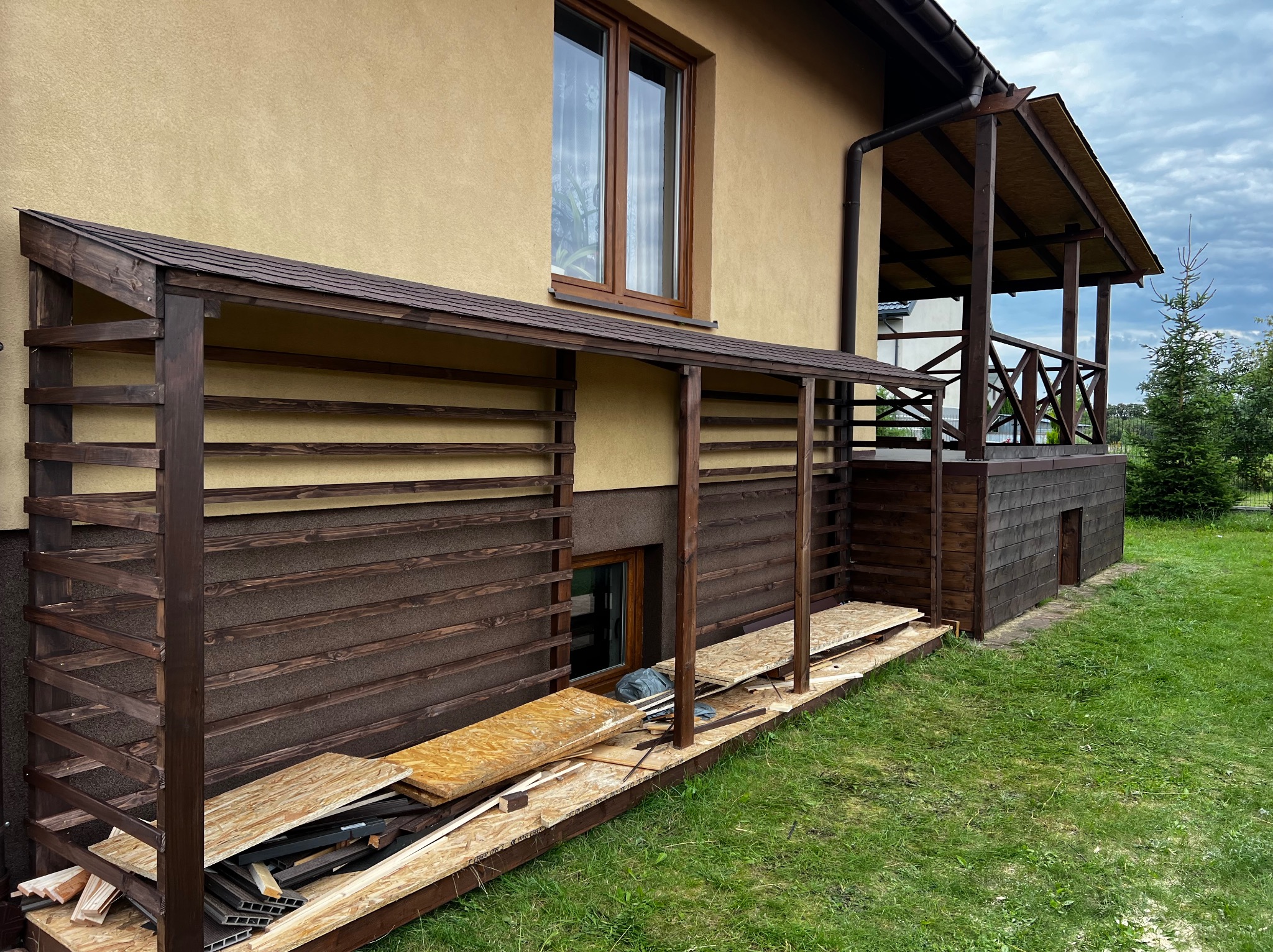 Drewniana konstrukcja zadaszenia przy budynku w trakcie budowy, widoczne deski i materiały budowlane. Częściowo zadaszony taras z balustradą w tle.