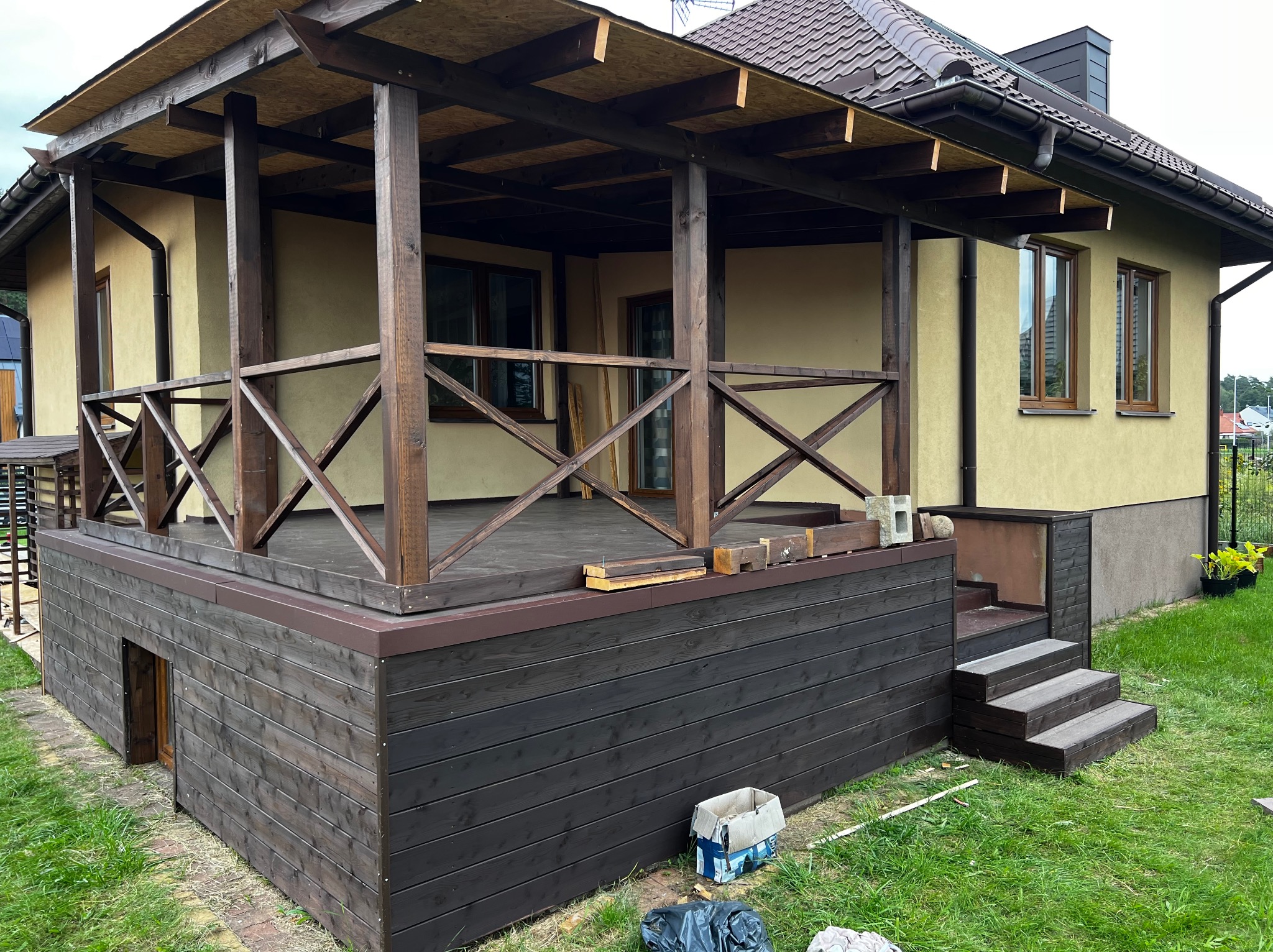 Drewniany taras z balustradą przy domu, z widocznymi schodami i elementami konstrukcyjnymi. Częściowo wykończony, z widocznymi narzędziami i materiałami budowlanymi.