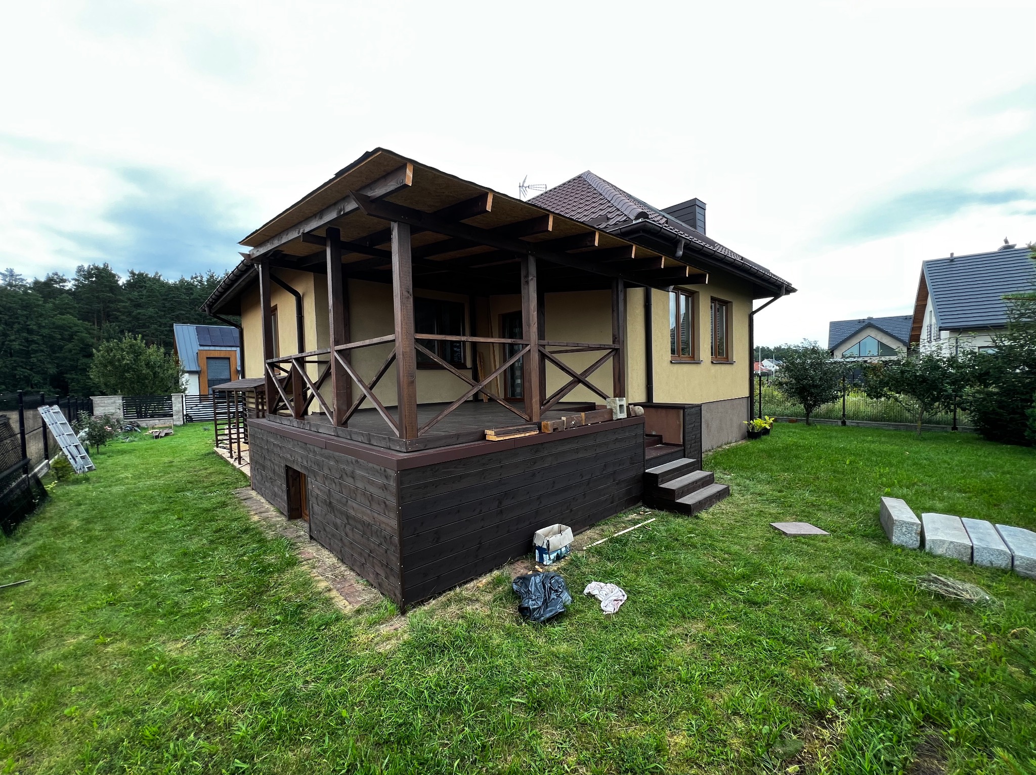 Drewniany taras z zadaszeniem w trakcie budowy przy domu jednorodzinnym. Widoczne materiały budowlane i narzędzia. Ujęcie z niskiej perspektywy, pochmurne niebo.