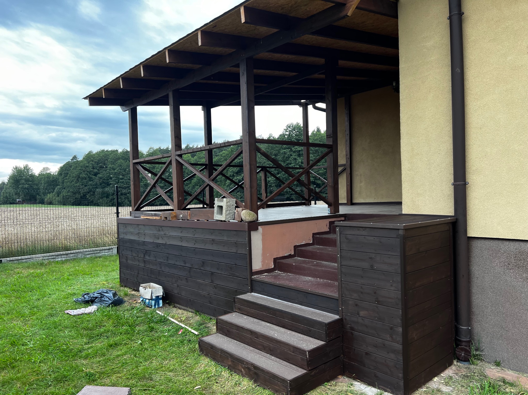 Drewniana pergola z zadaszeniem, brązowe elementy konstrukcyjne, schody wejściowe, widok na ogród i pole w tle, pochmurne niebo.