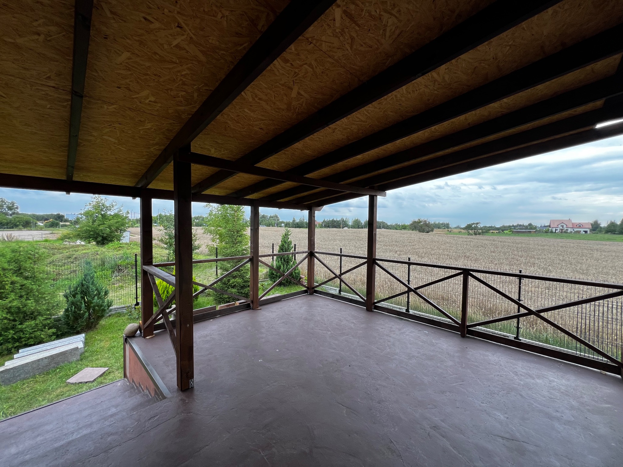 Zadaszony taras z drewnianą konstrukcją i widokiem na pole. Brązowa podłoga, drewniane barierki. Widok na krajobraz z zielenią i domem w oddali.
