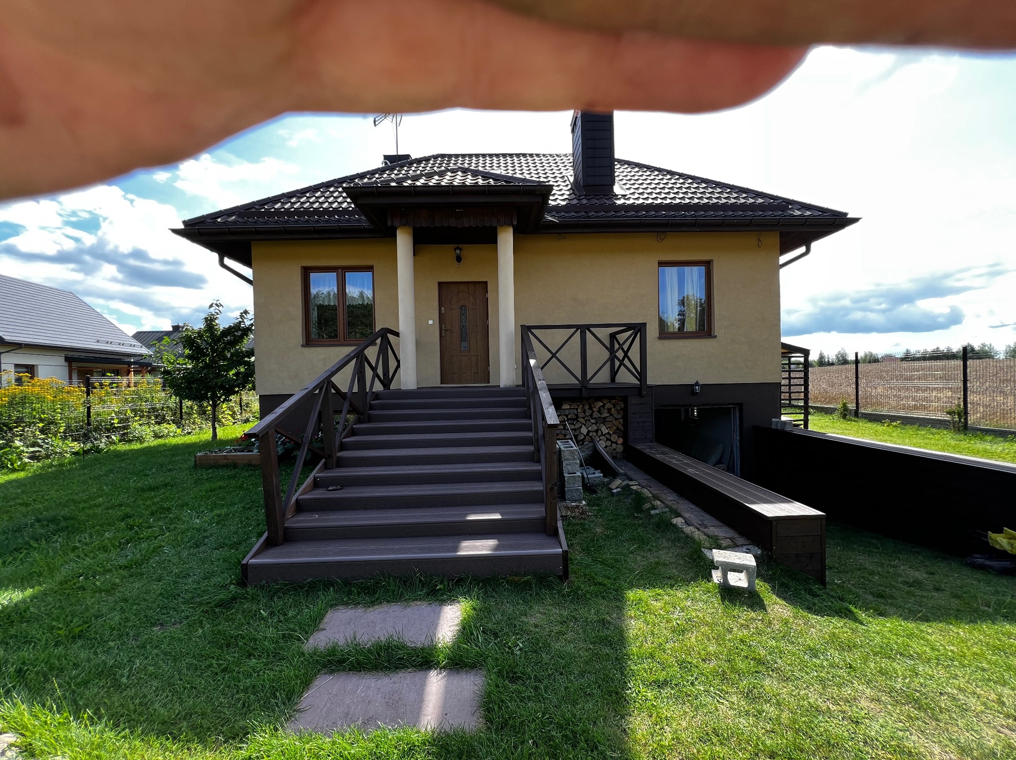 Dom z tarasem z drewnianymi schodami i balustradą. Widoczne wejście do piwnicy i elementy drewniane na elewacji. Ujęcie z poziomu trawnika.