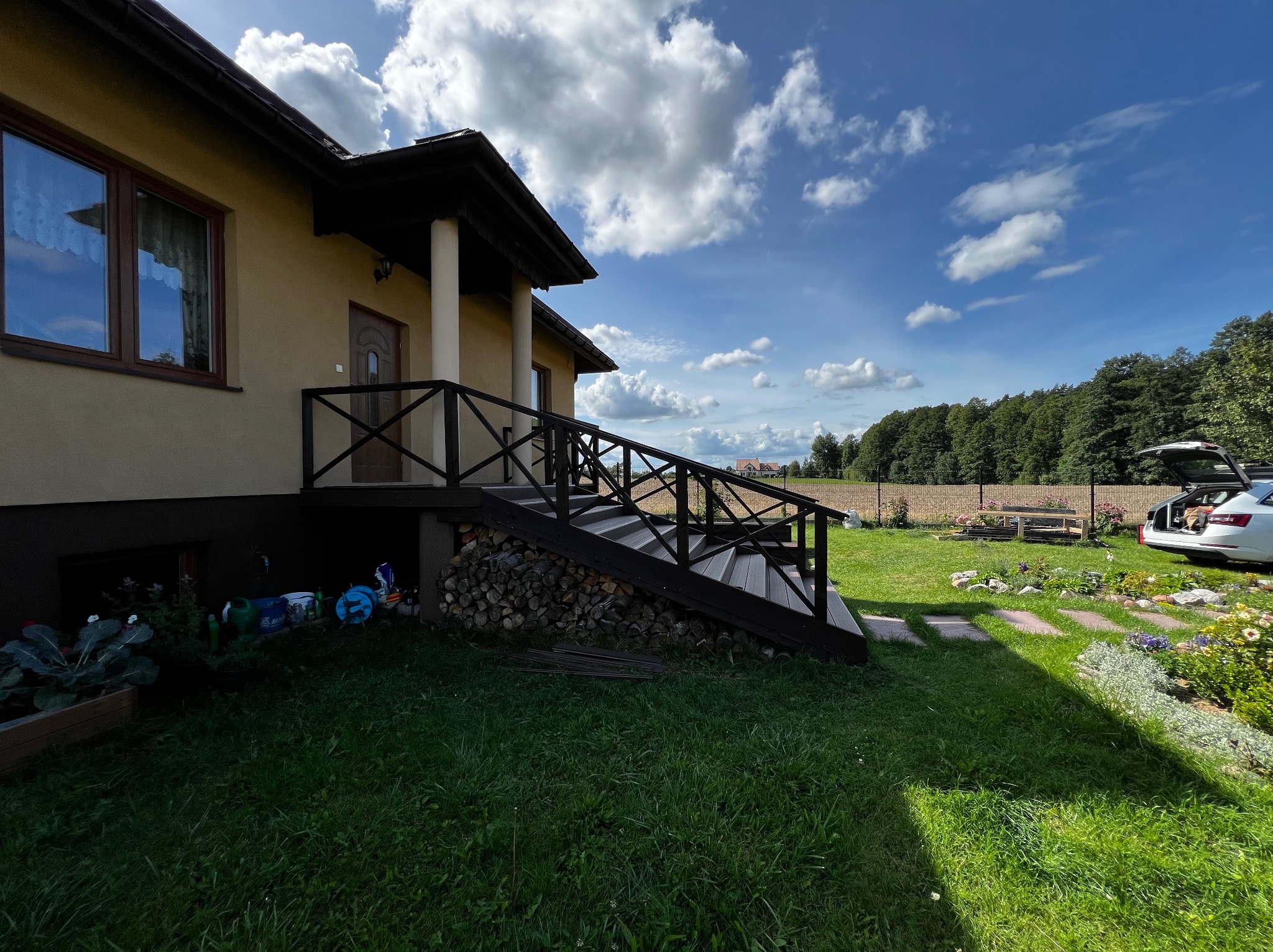 Drewniany taras z balustradą przy domu z kolumnami, schody z szarego materiału, ułożone drewno pod schodami, trawnik, błękitne niebo z chmurami w tle.