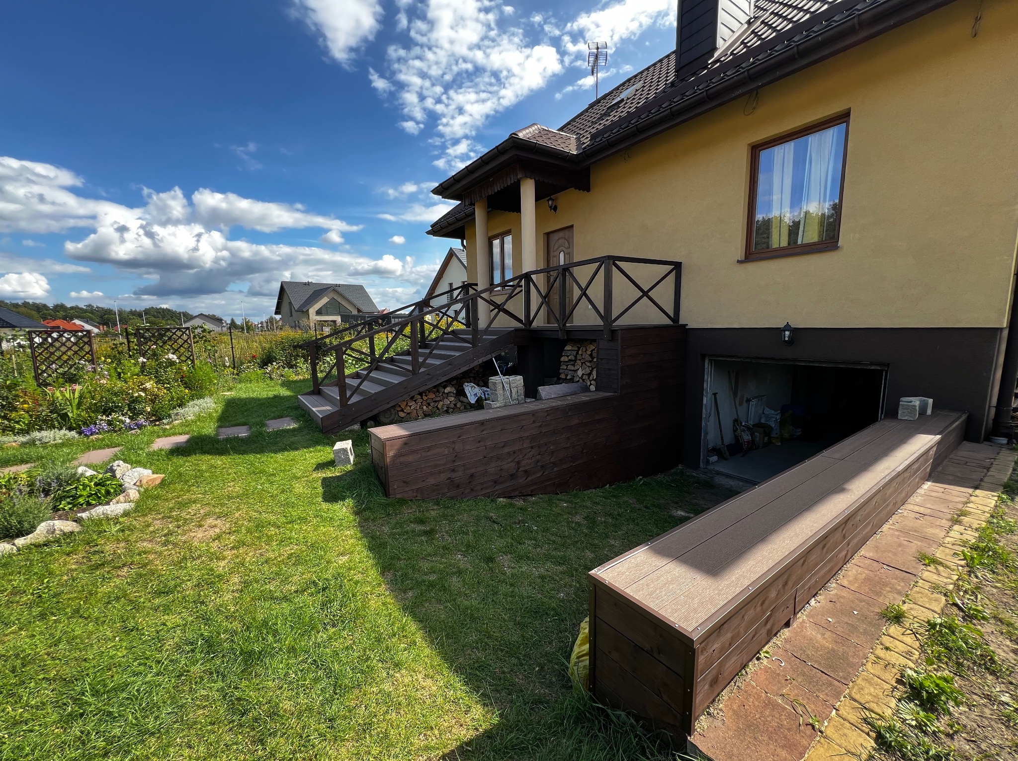 Dom z drewnianym tarasem i schodami, widok z ogrodu. Elementy drewniane w kolorze ciemnego brązu, kontrastują z jasną elewacją budynku. Błękitne niebo z chmurami w tle.