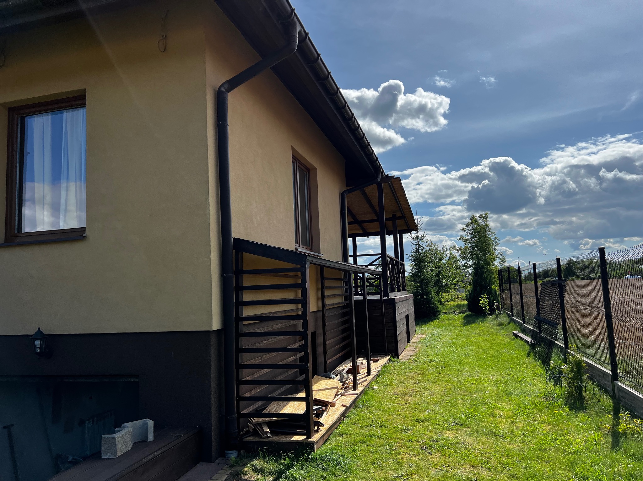 Dom z drewnianym tarasem i pergolą w trakcie budowy, widok z boku na tle zielonego trawnika i błękitnego nieba z chmurami. Widoczne materiały budowlane.