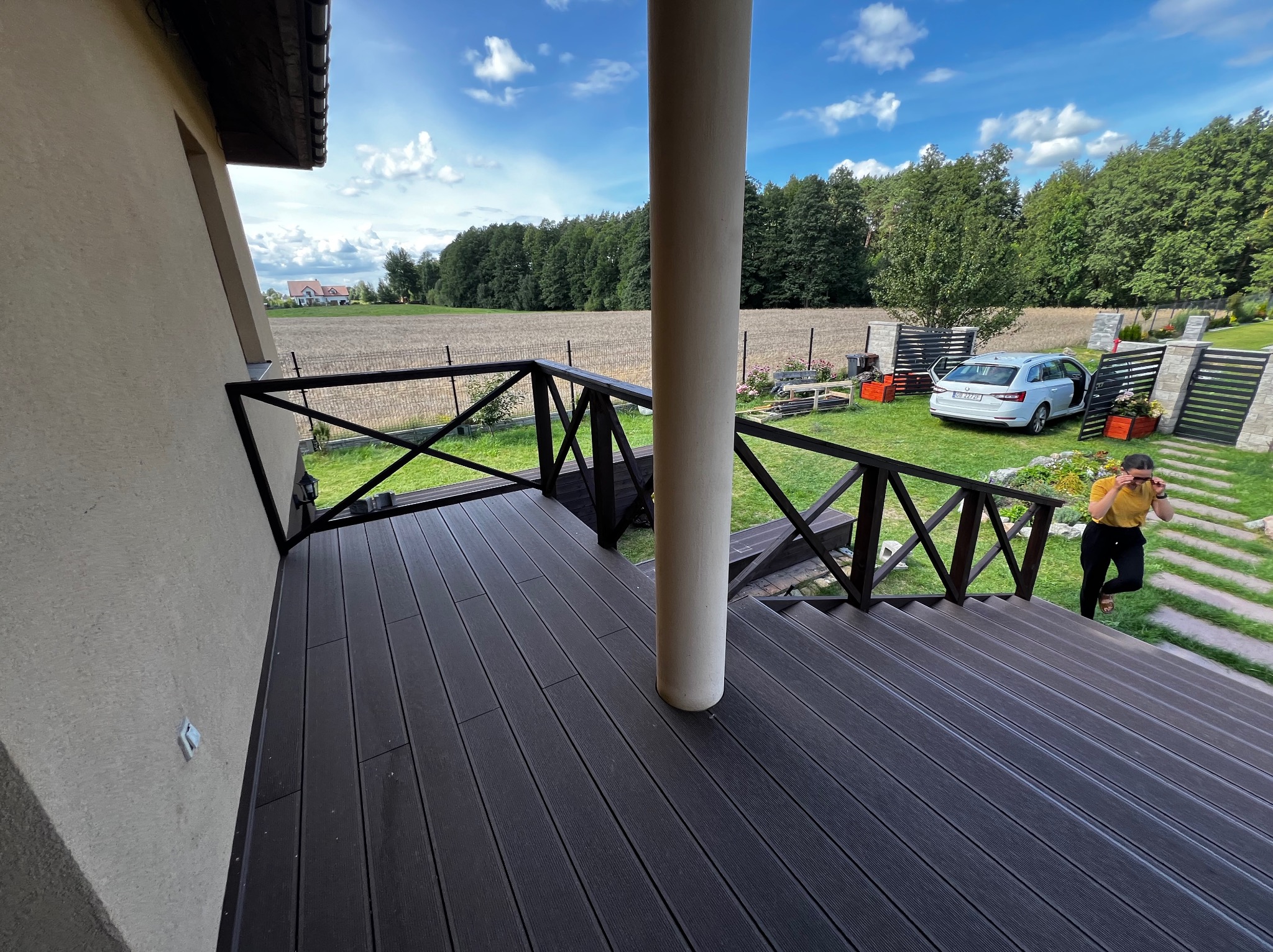 Nowoczesny taras z deski kompozytowej w kolorze ciemnego brązu, z czarną balustradą i widokiem na ogród. Na tarasie stoi biała kolumna, a w tle widać dom i samochód.