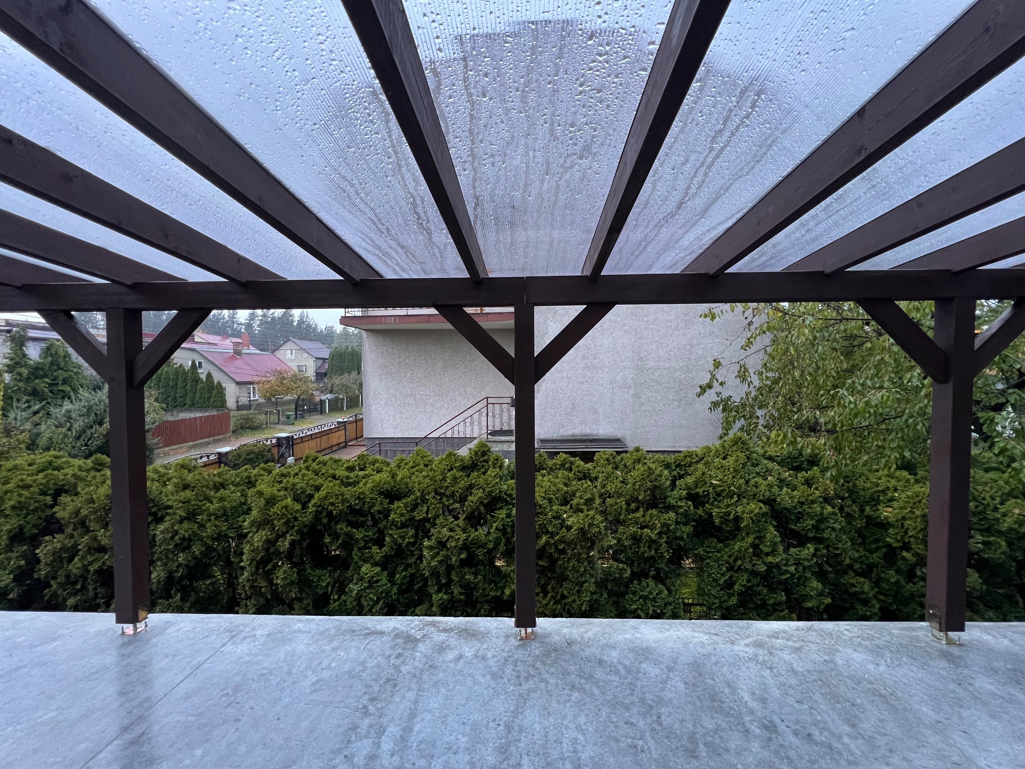 Drewniana pergola z przezroczystym zadaszeniem, widok z dołu na zewnątrz, krople deszczu na dachu, w tle zieleń i budynki mieszkalne.