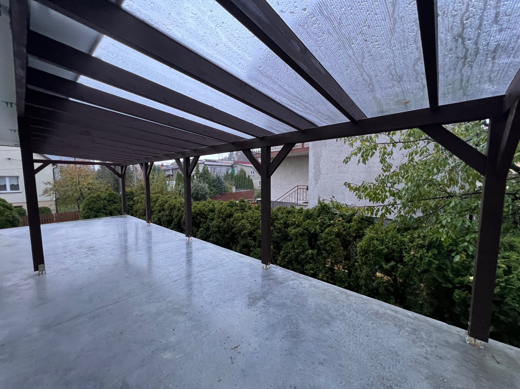 Zadaszenie tarasu z brązową konstrukcją drewnianą i przezroczystym dachem z tworzywa. Widok na ogród i budynki w tle. Lekko mokra nawierzchnia tarasu.