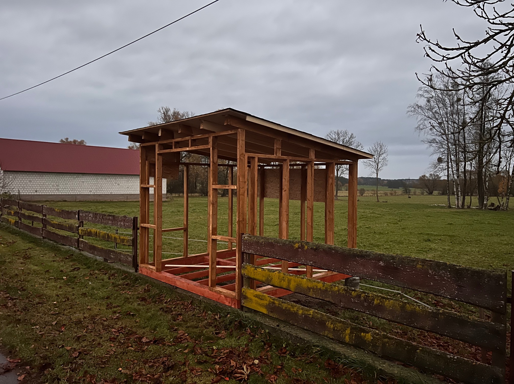 Drewniana konstrukcja szkieletowa z zadaszeniem w trakcie budowy na tle łąki i budynku. Widoczny fragment starego, drewnianego płotu z mchem.