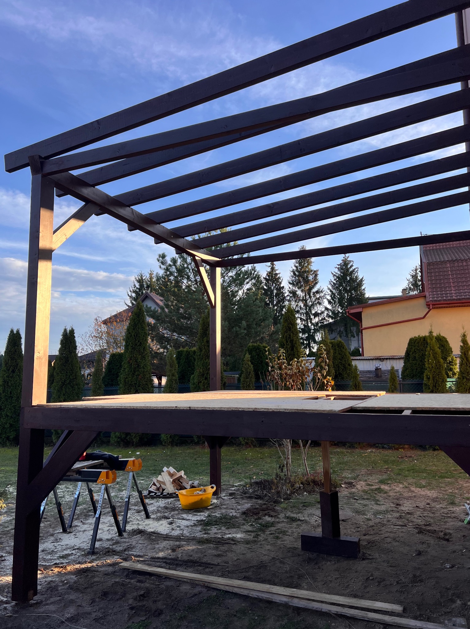 Wznoszenie drewnianej pergoli w ogrodzie, widoczne elementy konstrukcyjne i narzędzia. Ujęcie z dołu, w tle dom i drzewa, błękitne niebo.