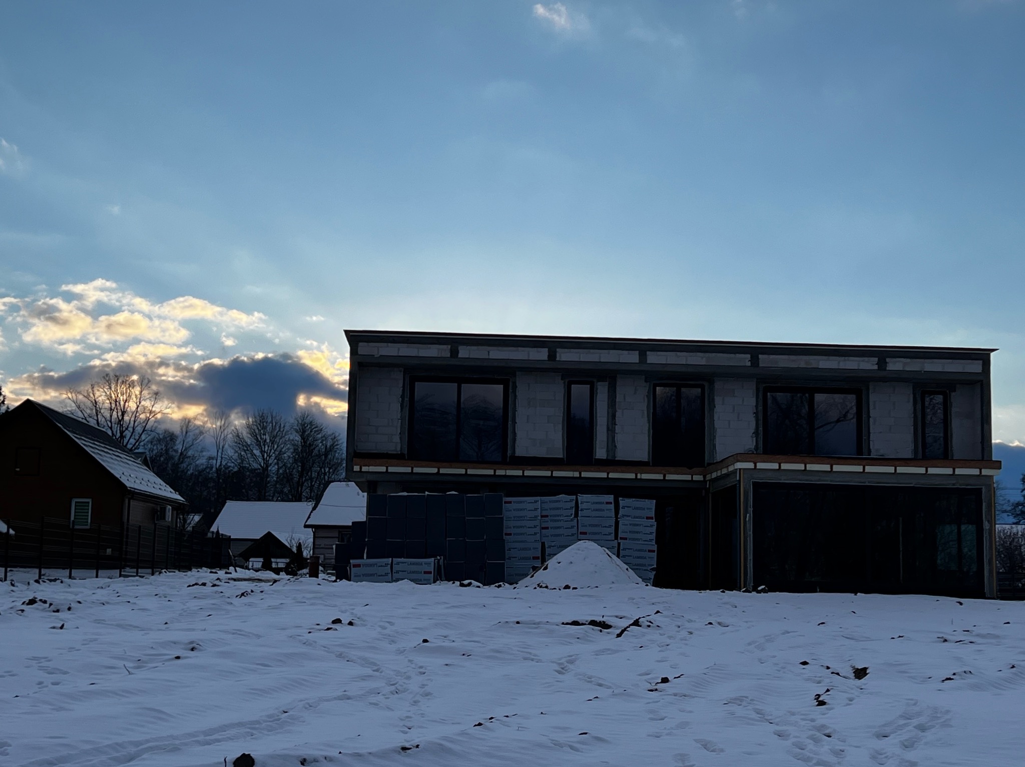 Surowy, dwukondygnacyjny budynek z pustaków w zimowym krajobrazie. Widoczne okna bez osłon, materiały budowlane i śnieg. Niebo z chmurami w tle.