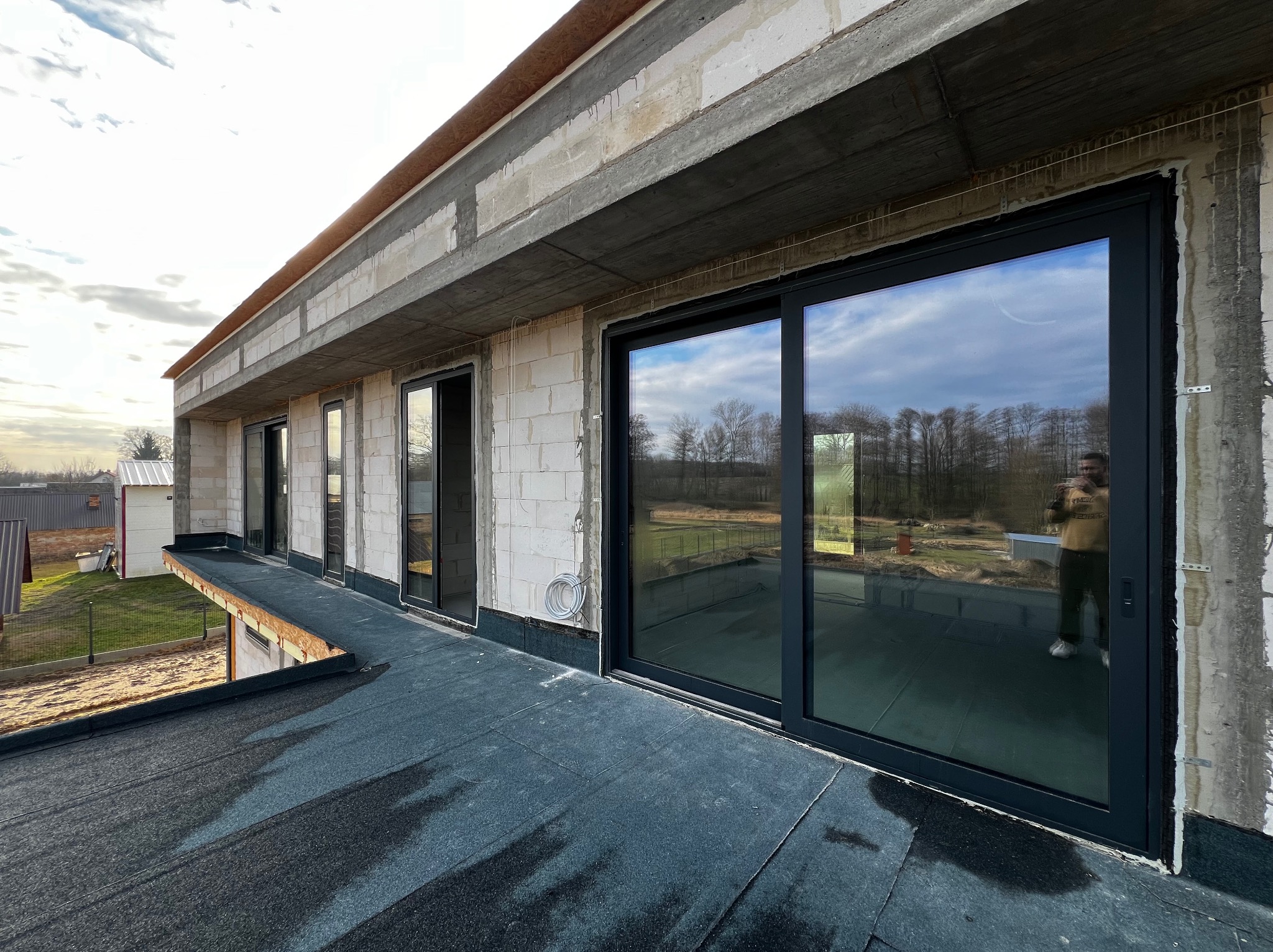 Montaż okien przesuwnych w surowym budynku z betonu komórkowego. Widoczne czarne ramy okienne i fragment dachu pokrytego papą. W tle drzewa i zabudowania.