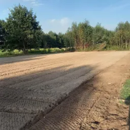 Przygotowane pole pod trawnik z rolki, widoczne ślady opon, drzewa w tle, błękitne niebo z chmurami.