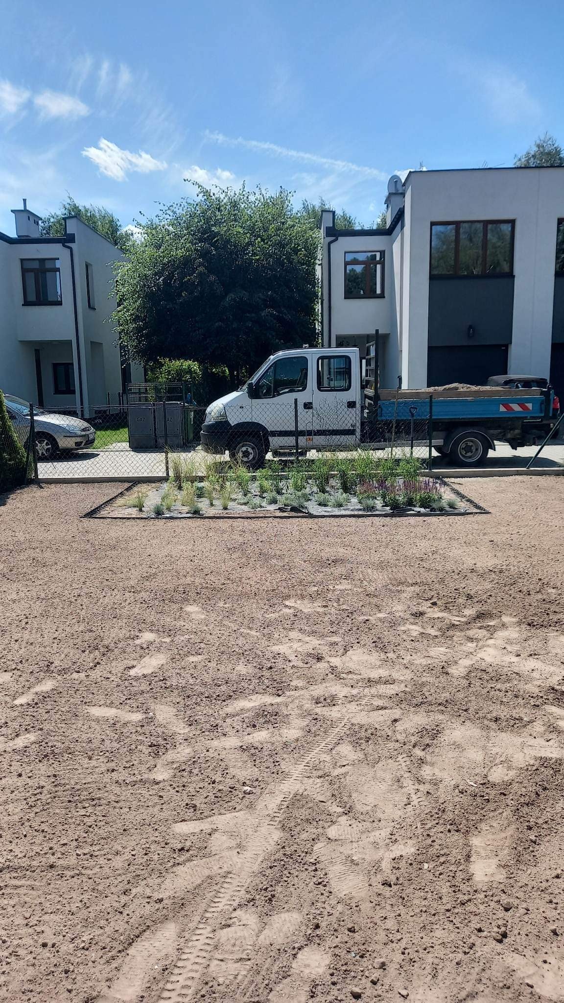 Przygotowany teren pod trawnik z widocznymi świeżo posadzonymi roślinami ozdobnymi, obok zaparkowany samochód dostawczy z przyczepą wypełnioną ziemią.