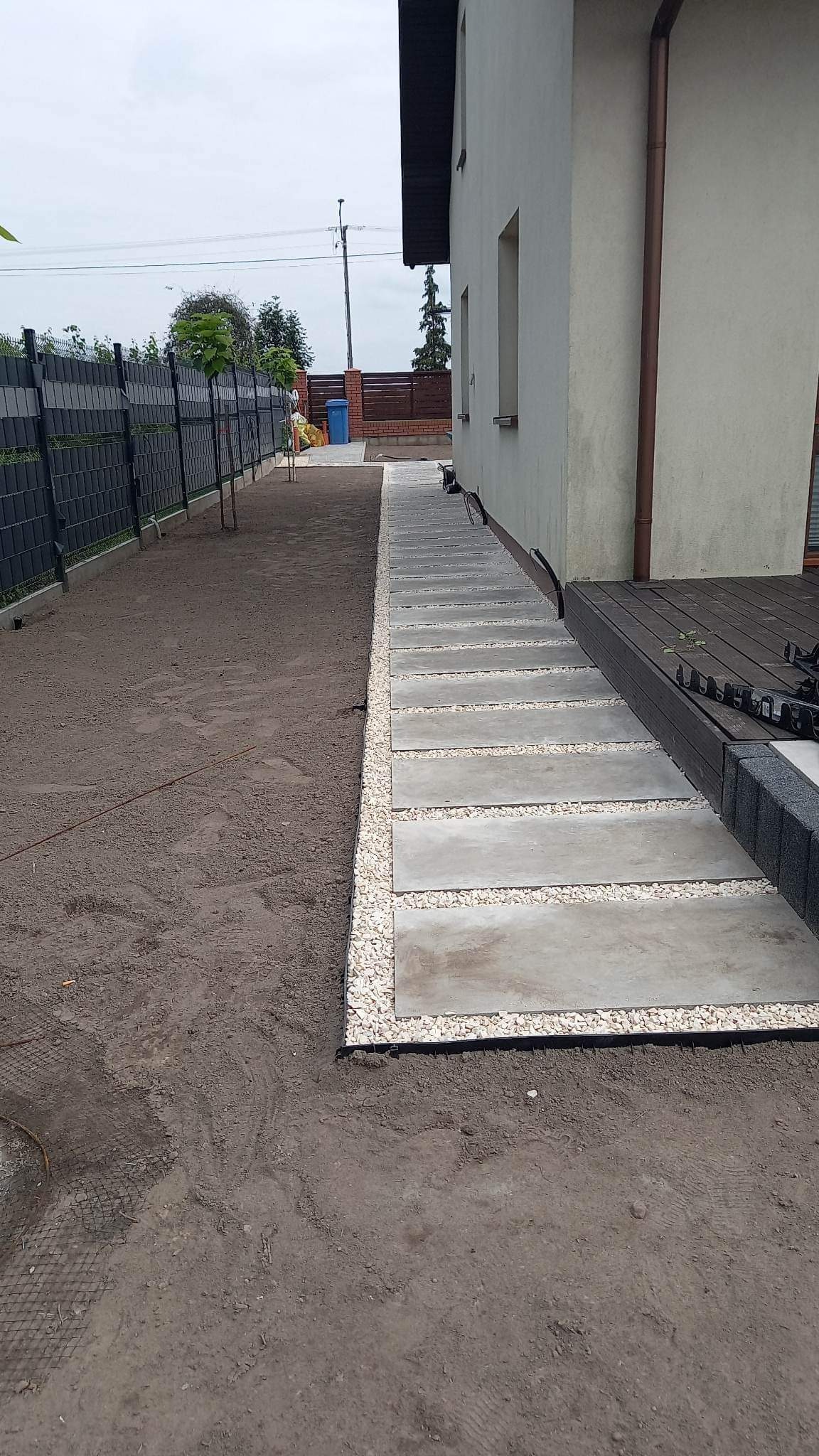 Ścieżka z betonowych płyt oddzielona od ziemi białym żwirem, prowadząca wzdłuż boku domu z brązową rynną i drewnianym tarasem.