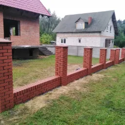 Ceglany murek ogrodzeniowy z filarami w trakcie budowy, widoczne dwa domy w tle, jeden w stanie surowym otwartym z czerwonej cegły, drugi otynkowany z szarym dachem.