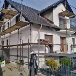 Dwupiętrowy dom w trakcie ocieplania, z rusztowaniami, widoczne płyty styropianowe na ścianach, pracownik w trakcie pracy, częściowo ukończona elewacja, widoczny czerwony samochód dostawczy.