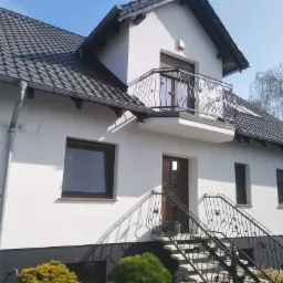 Dwukondygnacyjny dom murowany z białą elewacją, balkonem z kutą balustradą i ciemną dach&oacute;wką, widok z poziomu gruntu na fasadę i fragment ogrodu.