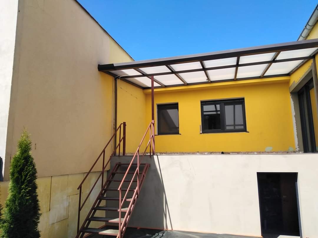 Żółta elewacja domu z czarnymi oknami i zadaszeniem, widoczne schody zewnętrzne z metalową barierką, fragment ocieplenia ściany z lewej strony.